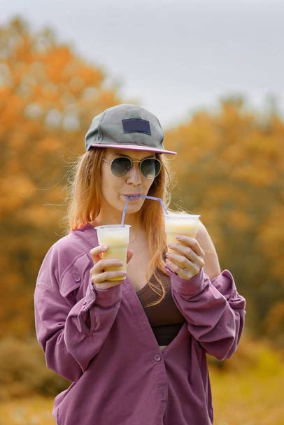 A girl in a purple shirt with purple lips drinks two milkshakes from tubules at the same time on an autumn day - Fotó, kép