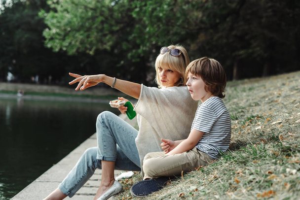 Mother and son sitting on the river bank in the park. Kid and his mom relaxing on pond shore in the grass. Woman points her hand on the lake. Family outside activities near water - Photo, Image