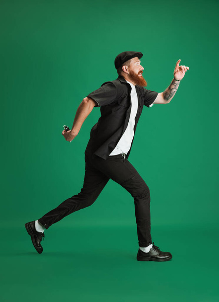 Portrait of stylish funny red-bearded man, barber or hair cutter running isolated on green studio background. Holding his cutting equipment. - Fotografie, Obrázek