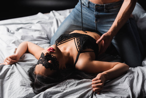 Sensual woman in lace mask lying on bed near boyfriend with spanking paddle on black background  - Фото, зображення