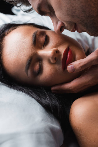 Mann berührt Kinn der brünetten Freundin mit geschlossenen Augen auf Bett auf schwarzem Hintergrund  - Foto, Bild