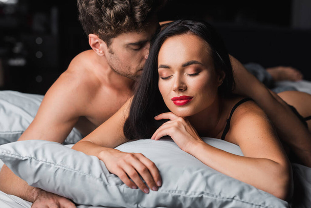 Hombre sin camisa abrazando a mujer sensual con labios rojos en la cama sobre fondo negro  - Foto, imagen