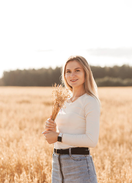 A young beautiful girl in a denim skirt walks through a wheat field on a sunny day - Zdjęcie, obraz