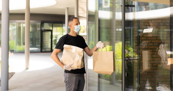 Entrega cara com máscara protetora e luvas segurando saco com mantimentos na frente de um prédio. - Foto, Imagem