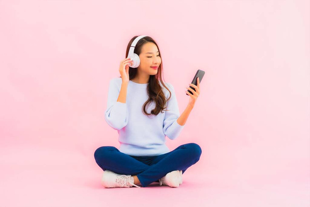 Retrato hermosa joven asiática mujer uso inteligente teléfono móvil con auriculares para escuchar música sobre fondo rosa - Foto, Imagen