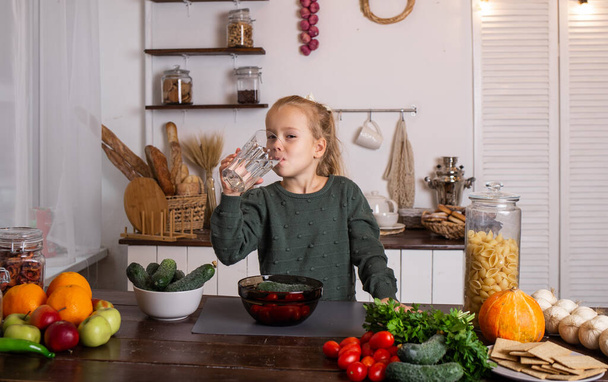 маленькая блондинка сидит за кухонным столом с овощами и фруктами и пьет воду из стакана - Фото, изображение