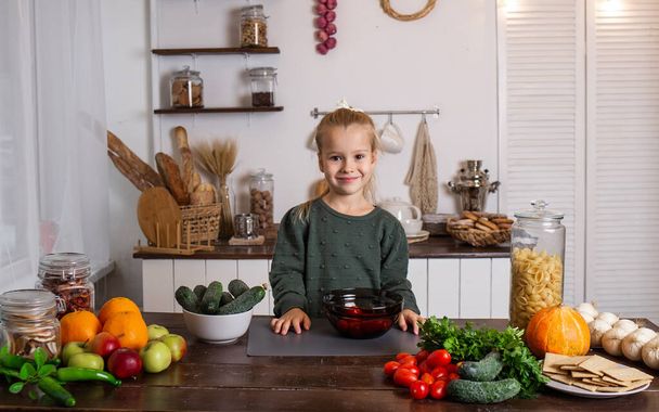 een klein blond meisje zit aan een tafel in de keuken met groenten en fruit - Foto, afbeelding
