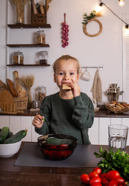pieni blondi tyttö istuu keittiön pöydässä vihannesten ja hedelmien kanssa ja syö leipää - Valokuva, kuva