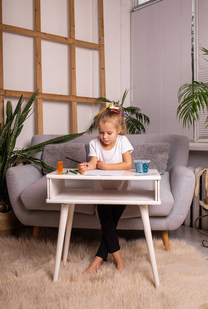 una niña rubia está sentada en una mesa y dibujando con lápices sobre papel - Foto, imagen
