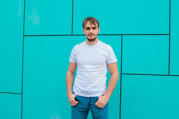 Beyaz tişörtlü genç bir adam mavi bir duvarın arkasında duruyor. Model. - Fotoğraf, Görsel