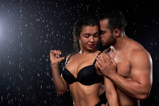 Portrait of passionate beautiful couple with athletic body under heavy rain on black - 写真・画像