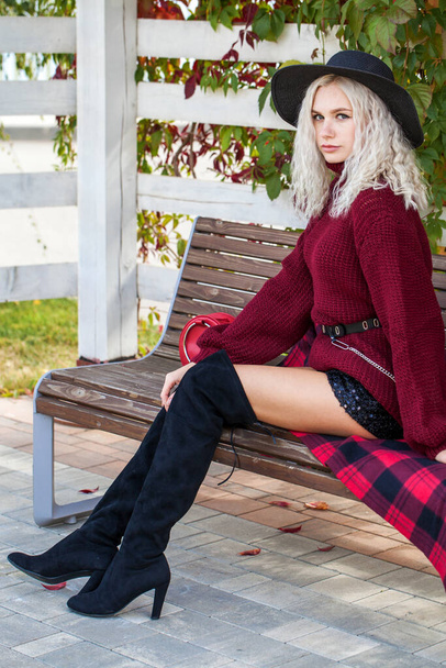 Young stylish girl in a burgundy sweater sits on a bench in an autumn park - Foto, immagini