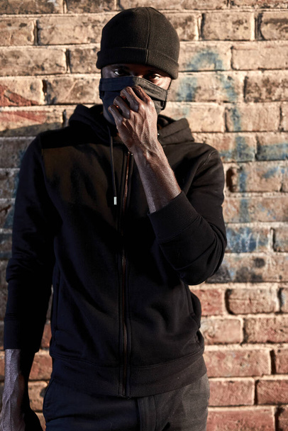 African american bandit man in mask standing by brick wall, hooligan in black wear - Фото, зображення