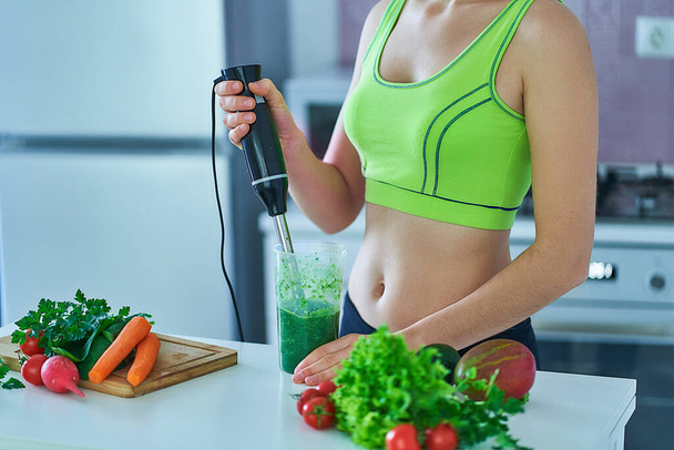 Sportive fitness woman in sportswear prepares a green smoothie using a hand blender at kitchen, healthy diet drinks - 写真・画像