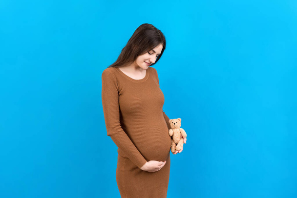 Mujer embarazada feliz sosteniendo un oso de peluche contra su vientre en fondo de color. La joven madre está esperando un bebé. Copiar espacio. - Foto, imagen