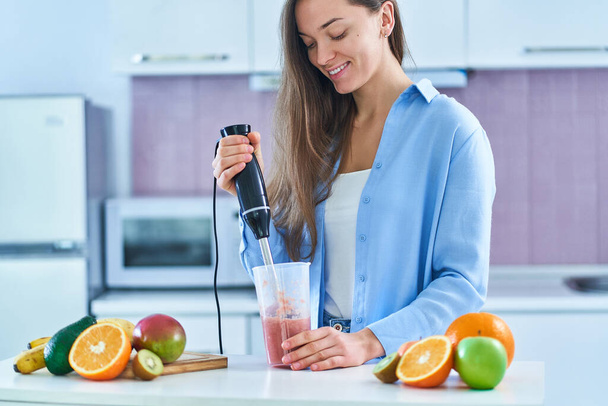 Happy cute female uses hand blender to mixing fresh organic fruits for prepare diet smoothie in the kitchen at home - Zdjęcie, obraz