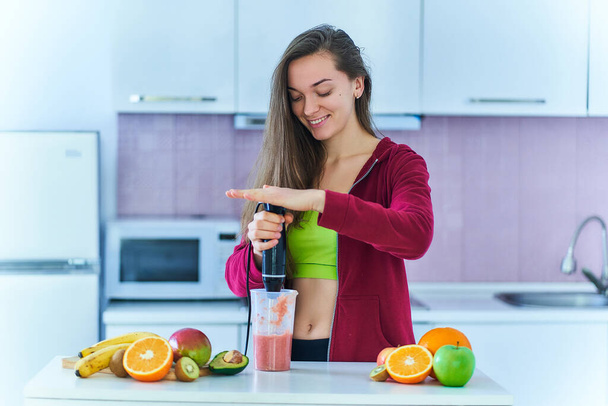 Happy joyful fitness female in sportswear prepares a fresh fruit smoothie using a hand blender at home in the kitchen. Vitamin diet drinks for losing weight and healthy eating  - Foto, Bild