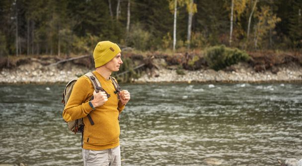 Viajante masculino está caminhando ao longo de um rio de montanha rápido com pedras enormes na costa contra o fundo de uma floresta de outono. Conceito de caminhadas e viagens - Foto, Imagem