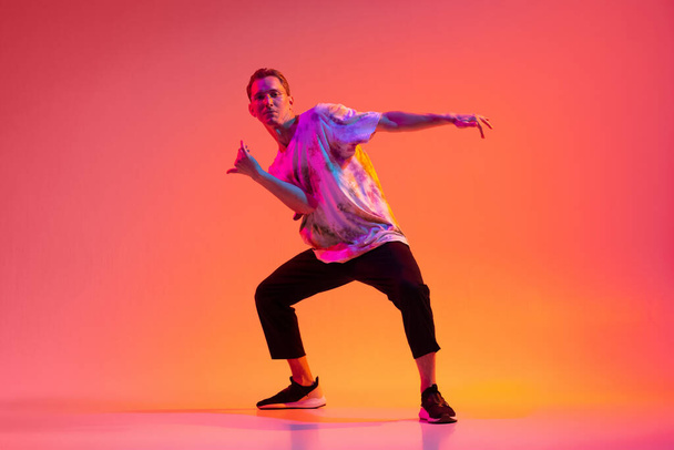 Portrait in motion of young caucasian man dancing hip-hop isolated over colorful gradient background in neon light. - Φωτογραφία, εικόνα