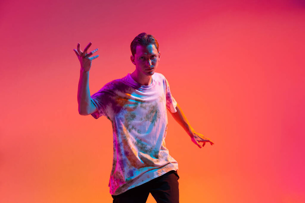 Ausgeschnittenes Porträt eines attraktiven jungen Mannes, der isoliert Hip-Hop-Tanz über einem Gradienten praktiziert, gelb-rosa Studiohintergrund in Neonlicht - Foto, Bild