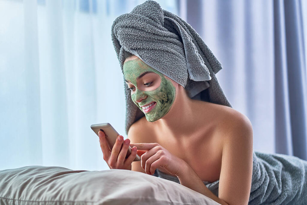 Cheerful happy female with green cleansing clay mask in bath towel and with smartphone is resting and relaxing after shower during spa day at home. Face skin care                                        - Fotografie, Obrázek