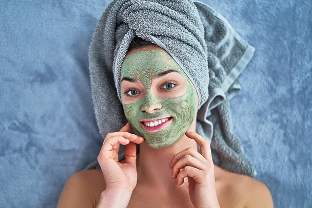 Happy woman with green cleansing clay mask in bath towel is resting and relaxing during spa day at home. Face skin care - Zdjęcie, obraz