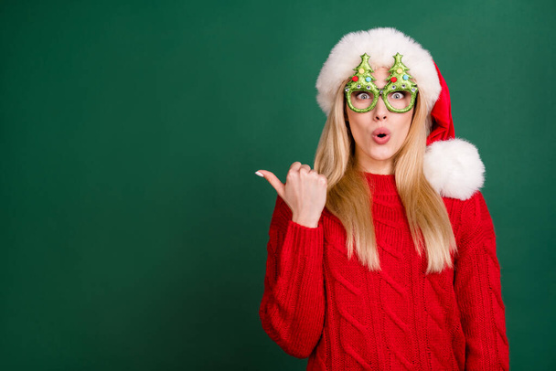 Photo portrait blonde woman in santa hat amazed showing empty space on thumb isolated green color background - 写真・画像