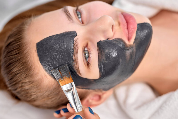 Female relaxing with charcoal facial mask, cropped professional beautician applying on face - Фото, изображение