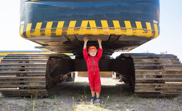4-year-old boy in a red uniform and cap speaks on a cell phone at a construction site, plays with toy cars on the track of a large excavator. Positive, interesting childhood, games in the profession - Valokuva, kuva