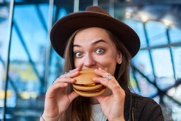 Happy joyful stylish hipster hungry woman with surprised wide eyes eats burger in fast food restaurant - Zdjęcie, obraz