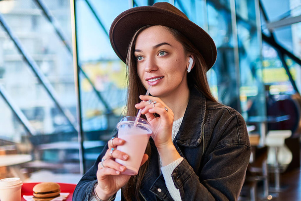 Portrait of stylish casual hipster drinking woman with sweet milkshake in hat with wireless earphones during relaxing in fast food restaurant. Modern people lifestyle - Φωτογραφία, εικόνα