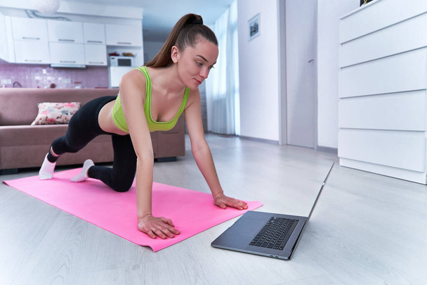 Mujer atlética fuerte en ropa deportiva durante la observación de entrenamiento de fitness en un ordenador portátil y hacer ejercicios de fitness en la estera. Entrenamiento en línea en el gimnasio en casa     - Foto, Imagen