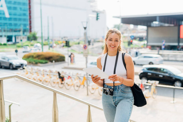 Mujer joven caucásica caminando al aire libre usando auriculares y tabletas trabajando como freelance - Foto, imagen