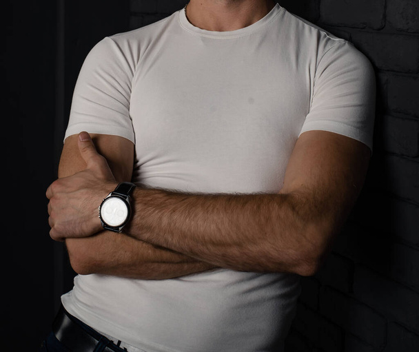 Unrecognizable man in a white T-shirt with a watch on his hand on a dark background. - Фото, изображение