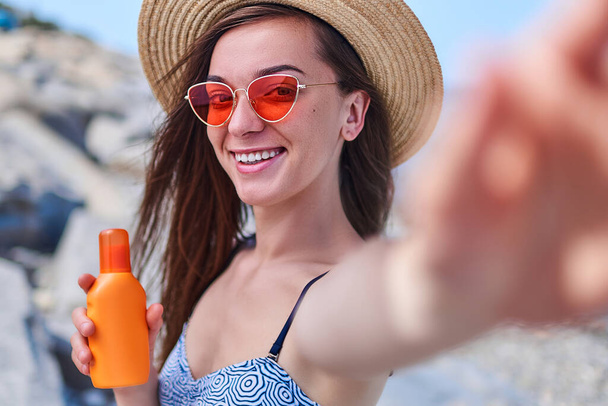 Portrait of a happy smiling woman in a swimsuit, straw hat and bright red glasses with a bottle of sun block cream during sunbathing by the sea in sunny weather in the summertime - Photo, image