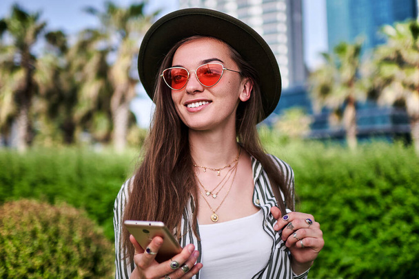 Stylish carefree young hipster woman wearing hat and bright red glasses using phone outdoors          - Foto, Imagem