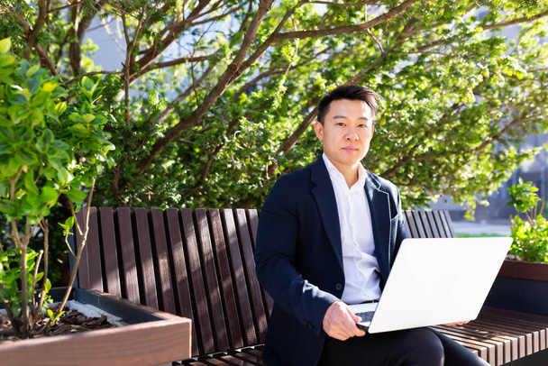 ernster asiatischer Mann im Business-Anzug arbeitet am Laptop und sitzt tagsüber auf der Bank und schaut in die Kamera - Foto, Bild