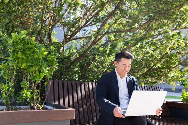 young asian business man or freelancer sitting on bench and working with laptop in city park on modern urban street background outdoors downtown Male enjoy outside Focused entrepreneur browsing typing - Zdjęcie, obraz