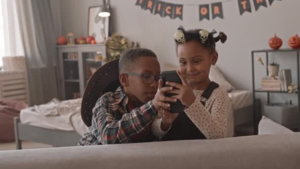 Slowmo shot de mignonne fille afro-américaine de 5 ans et son frère aîné s'amuser à regarder des vidéos ou à jouer à des jeux sur smartphone pendant la fête d'Halloween à la maison - Séquence, vidéo