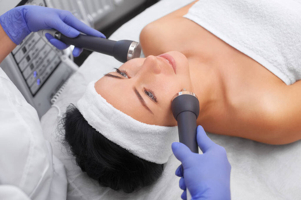 Le cosmétologue fait de la procédure un nettoyage ultrasonique de la peau du visage d'une belle jeune femme dans un salon de beauté.. - Photo, image