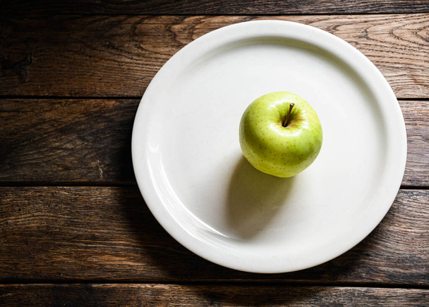 Одне зелене яблуко на білій керамічній тарілці, на дерев'яному столі
. - Фото, зображення