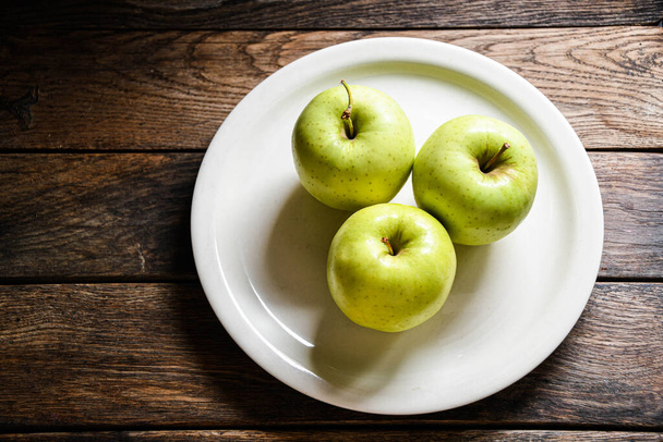Three green apples on a white ceramic plate, on a wooden table. - Φωτογραφία, εικόνα
