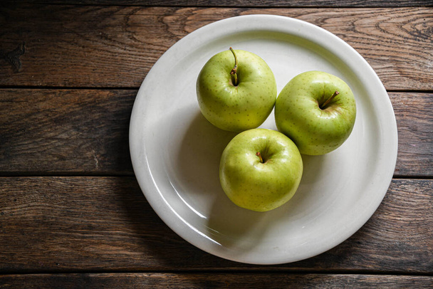 Три зелених яблука на білій керамічній тарілці, на дерев'яному столі
. - Фото, зображення