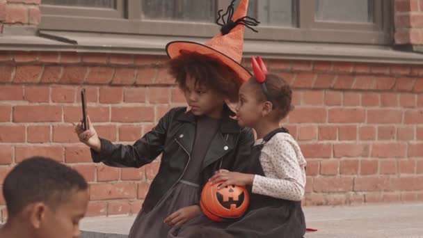 Lento disparo de lindas niñas afroamericanas de 5 y 8 años de edad con disfraces de Halloween sentadas al aire libre, comiendo dulces de cubos de calabaza y tomando retrato de selfie en el teléfono inteligente - Metraje, vídeo