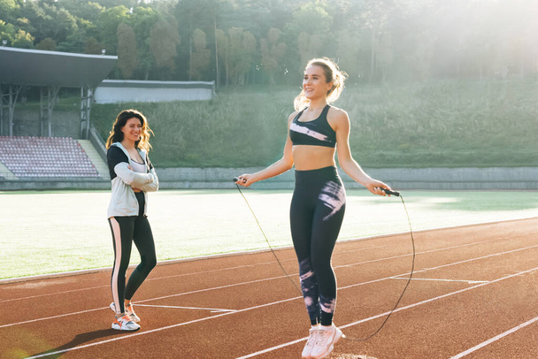 Sporty female with a good figure jumps rope on sports track of stadium. Athletic woman with personal instructor skipping rope as part of her fitness workout. Exercising strength cardio and power - Photo, image