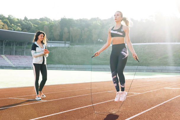 Sporty female with a good figure jumps rope on sports track of stadium. Athletic woman with personal trainer jumping rope as part of her fitness workout. Exercising strength cardio and power - Photo, image