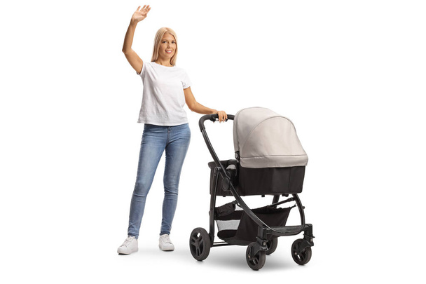 Full length portrait of a young casual mother in jeans with a pushchair waving at camera isolated on white background - Foto, Imagem