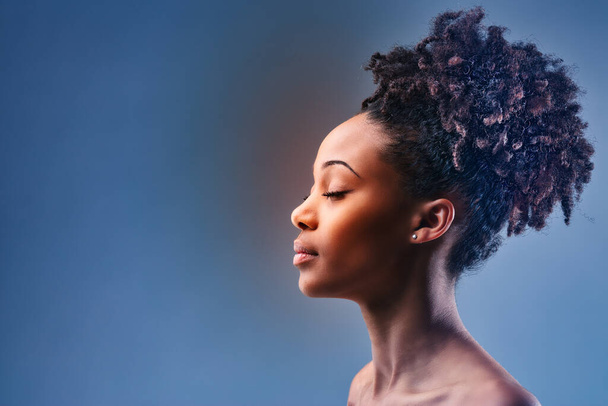 Perfil de cabeça e ombros retrato de uma jovem mulher negra posando com os olhos fechados, ombros nus e seu lindo cabelo encaracolado amarrado na parte superior da cabeça sobre azul com copyspace - Foto, Imagem