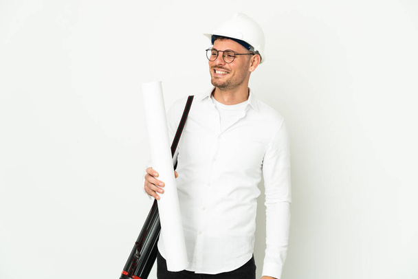 Jeune architecte homme avec casque et tenant des plans isolés sur fond blanc regardant sur le côté et souriant - Photo, image