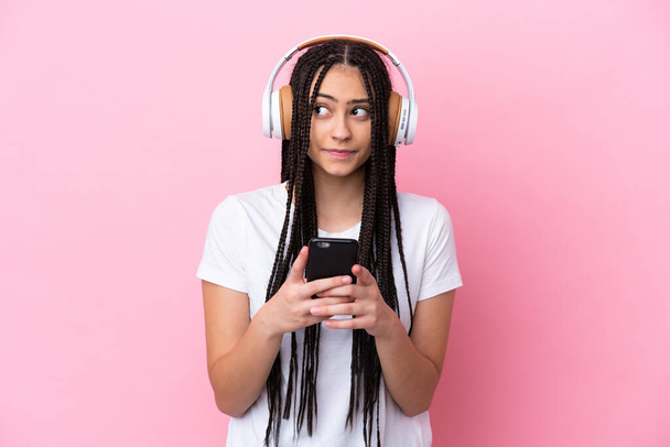 Девочка-подросток с косичками на изолированном розовом фоне слушает музыку с мобильного телефона и думает - Фото, изображение